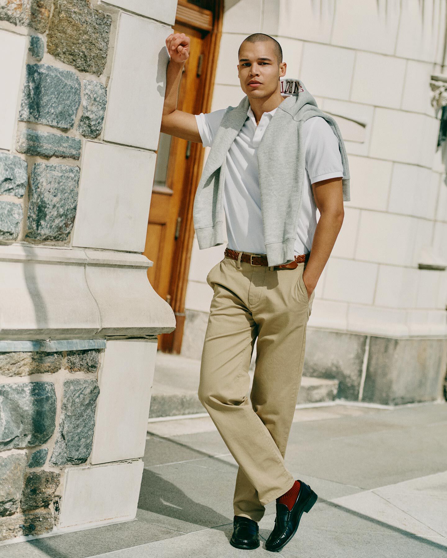 A man in a white polo and beige Gant chinos stands by a rustic stone wall and wooden door in soft, natural light.